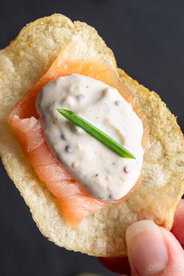 A hand holding a potato chip canapé topped with a slice of smoked salmon, a dollop of creamy sauce, and a small piece of chive on top.