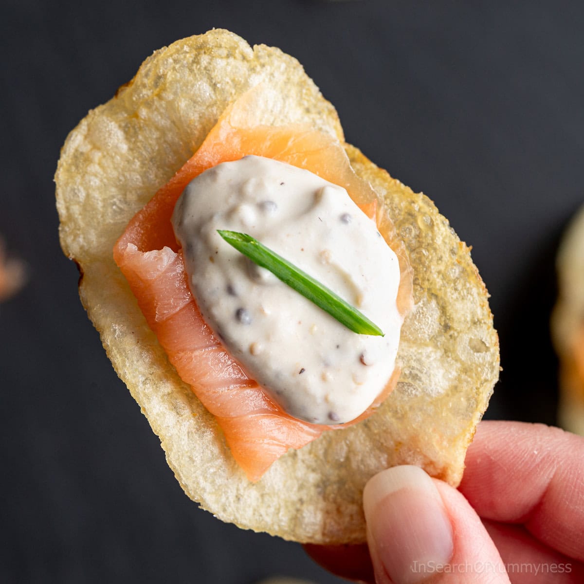 A hand holding a potato chip canapé topped with a slice of smoked salmon, a dollop of creamy sauce, and a small piece of chive on top.