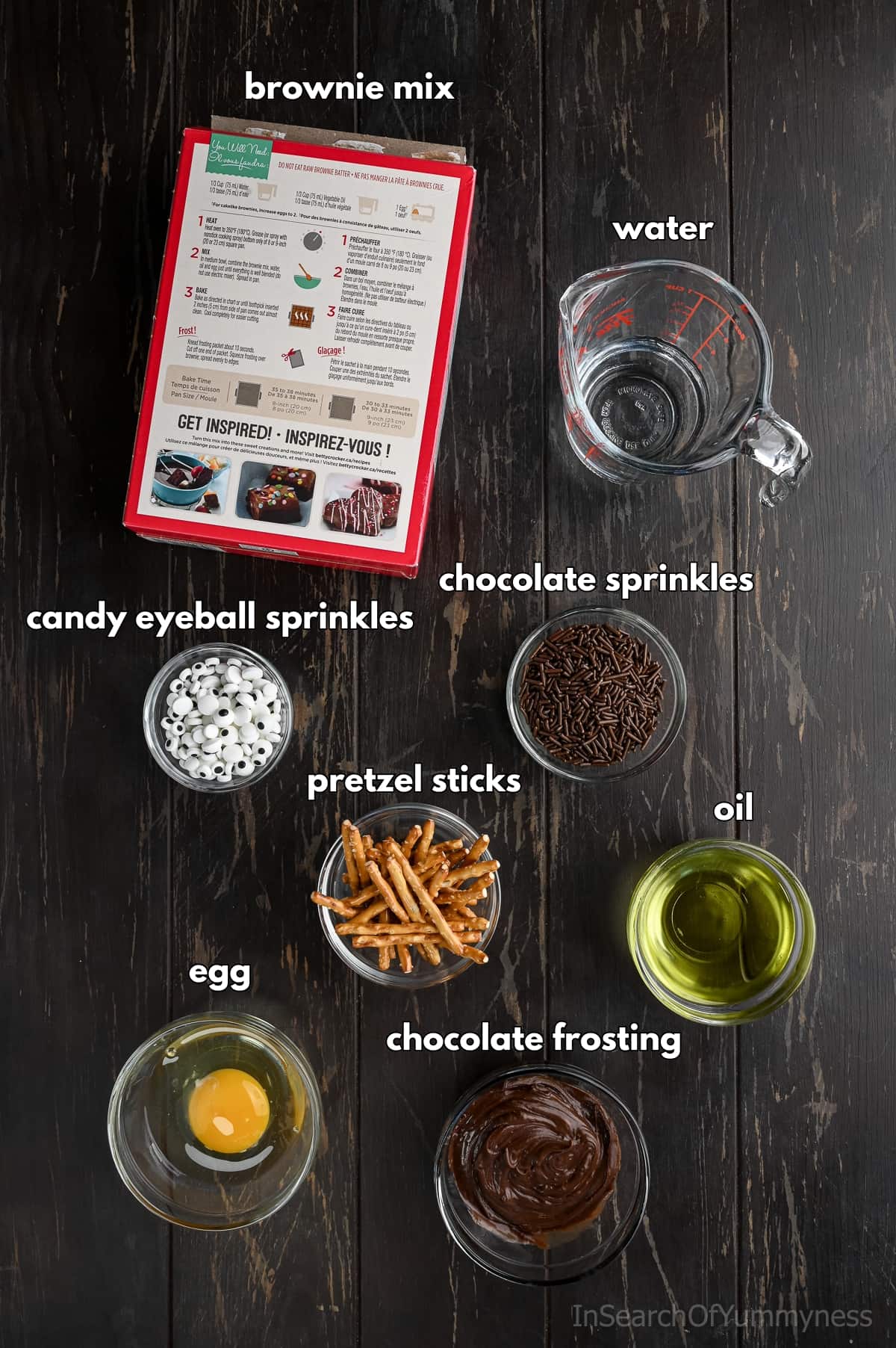 Ingredients for making Halloween spider brownies arranged on a dark wooden table. Includes a box of brownie mix, a glass measuring cup with water, a bowl of oil, a bowl with a cracked egg, pretzel sticks, candy eyeball sprinkles, chocolate sprinkles, and chocolate frosting.
