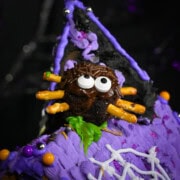 A decorated spider brownie with pretzel stick legs, chocolate frosting, candy eyeball sprinkles, and chocolate sprinkles sits on top of a purple icing-covered Halloween gingerbread house.