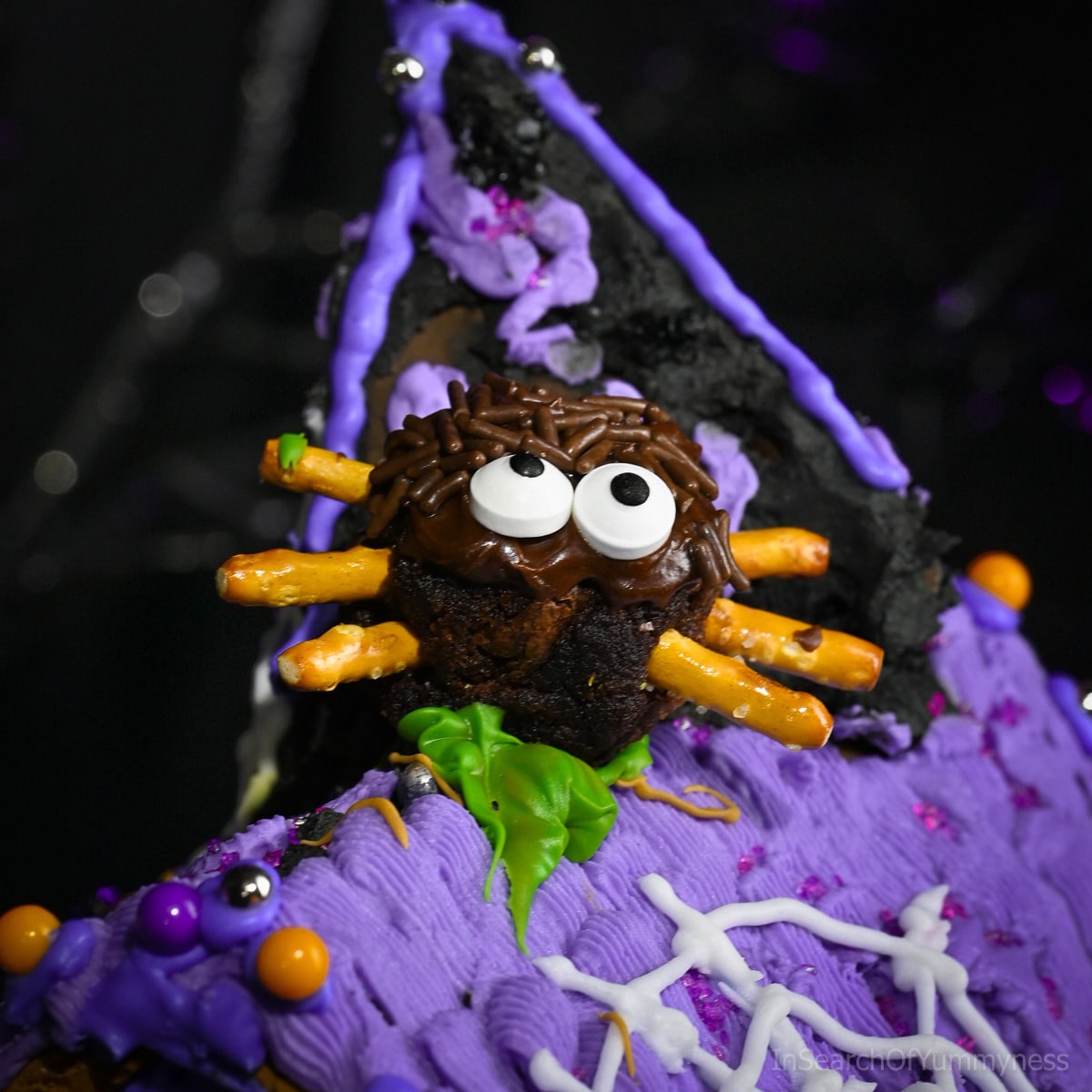 A decorated spider brownie with pretzel stick legs, chocolate frosting, candy eyeball sprinkles, and chocolate sprinkles sits on top of a purple icing-covered Halloween gingerbread house.