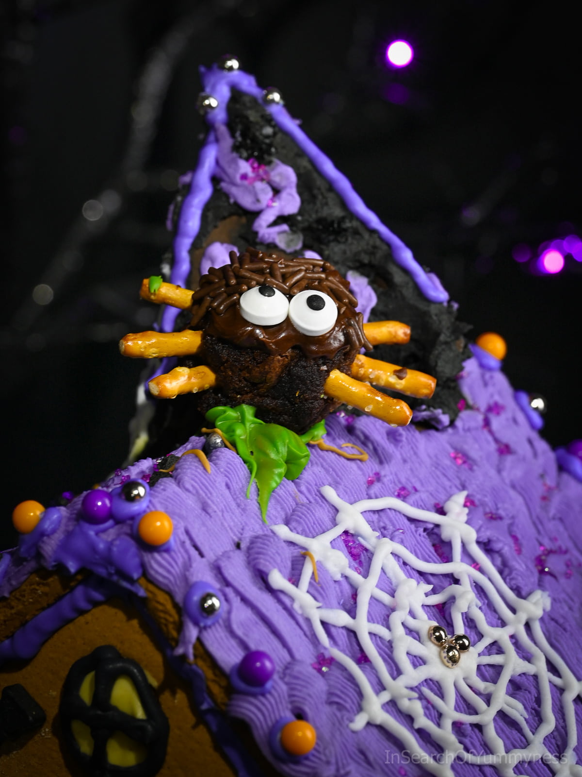 A decorated spider brownie with pretzel stick legs, chocolate frosting, candy eyeball sprinkles, and chocolate sprinkles sits on top of a purple icing-covered Halloween gingerbread house.