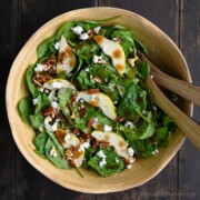 A wooden bowl filled with spinach pear salad topped with crumbled goat cheese, toasted pecans, and balsamic vinaigrette, with salad tongs resting on the side.