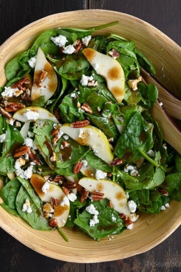 A wooden bowl filled with spinach pear salad topped with crumbled goat cheese, toasted pecans, and balsamic vinaigrette, with salad tongs resting on the side.
