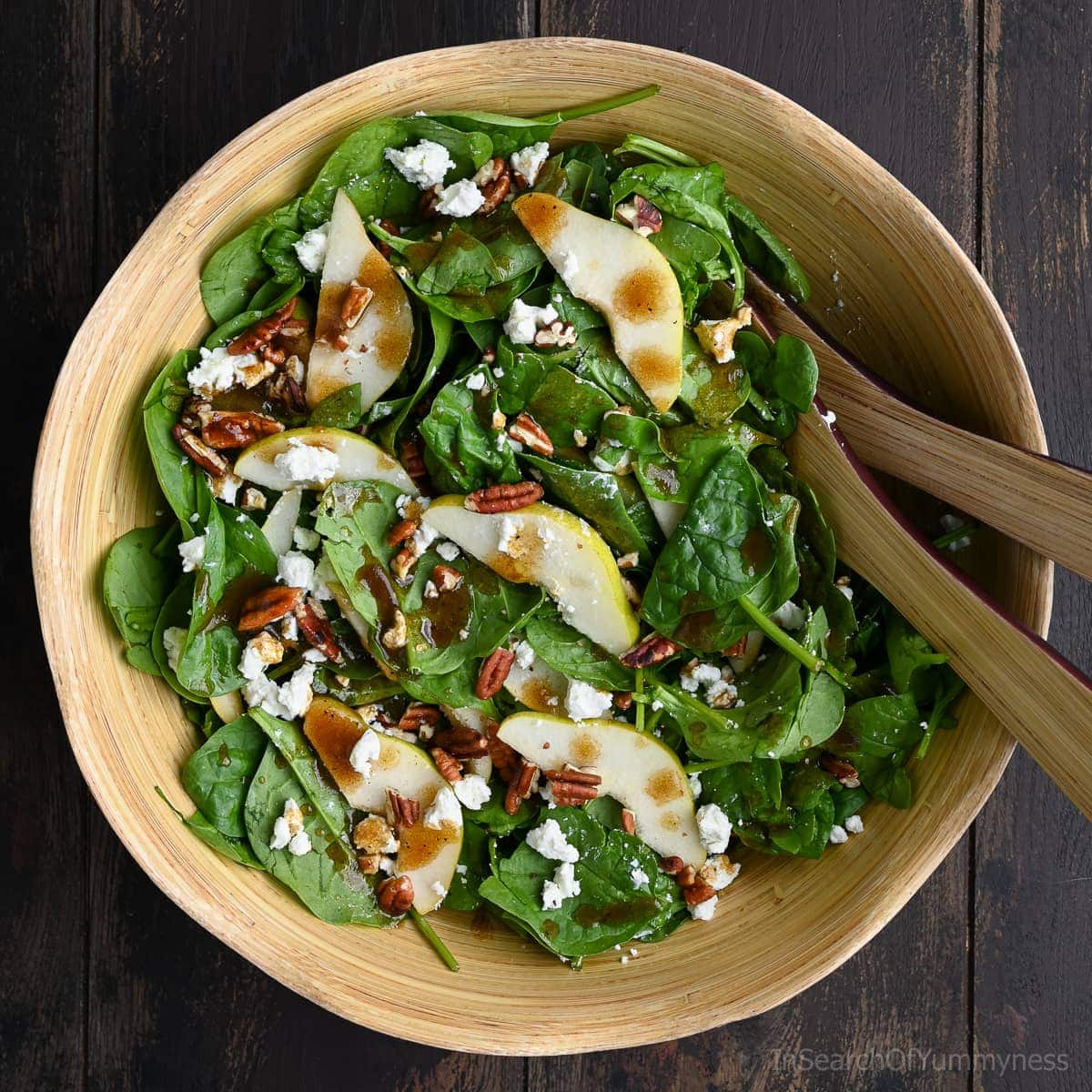 A wooden bowl filled with spinach pear salad topped with crumbled goat cheese, toasted pecans, and balsamic vinaigrette, with salad tongs resting on the side.