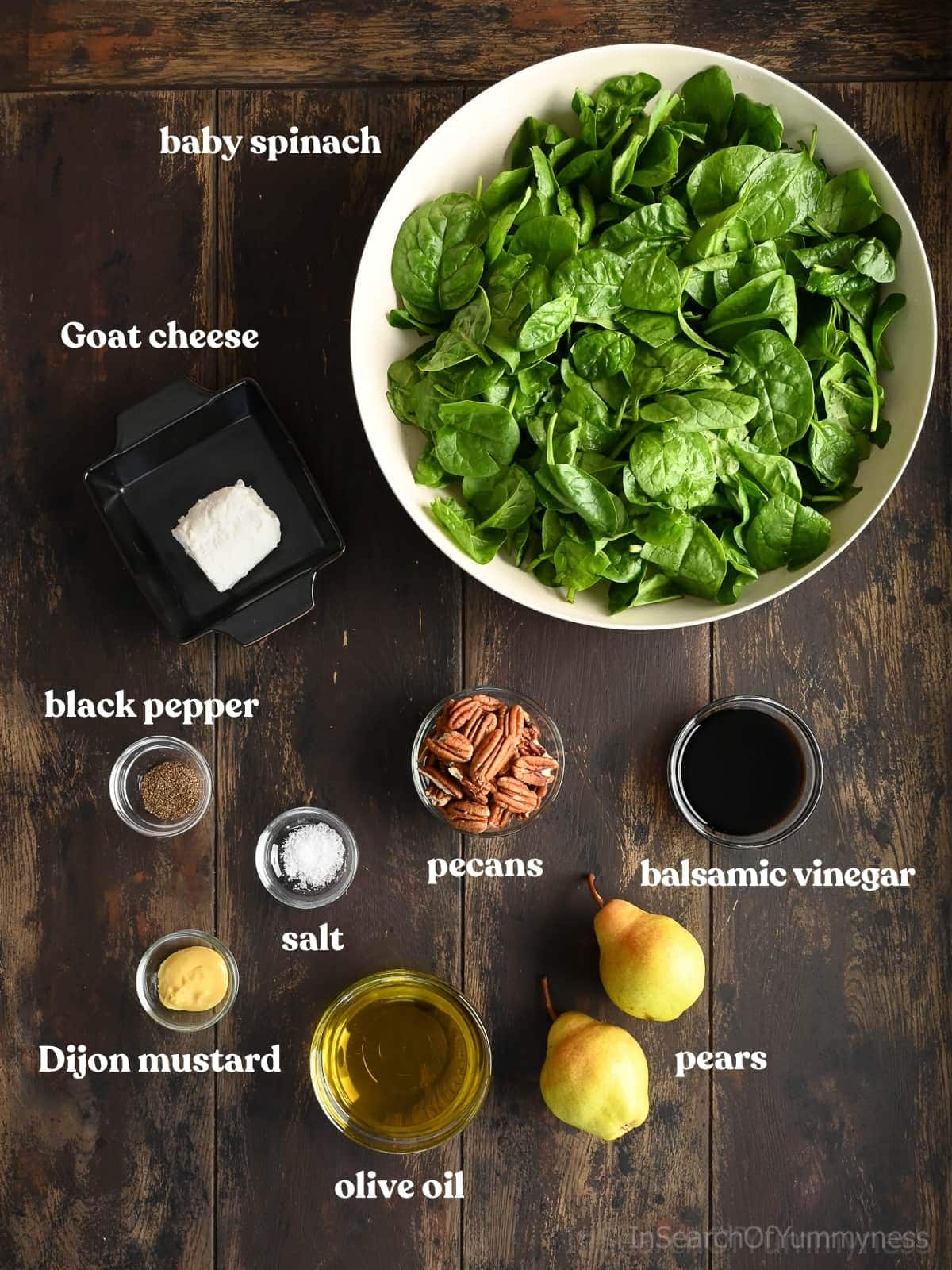 Overhead view of labeled ingredients for a spinach pear salad, including baby spinach, goat cheese, black pepper, salt, Dijon mustard, olive oil, pecans, balsamic vinegar, and fresh pears arranged on a dark wooden surface.
