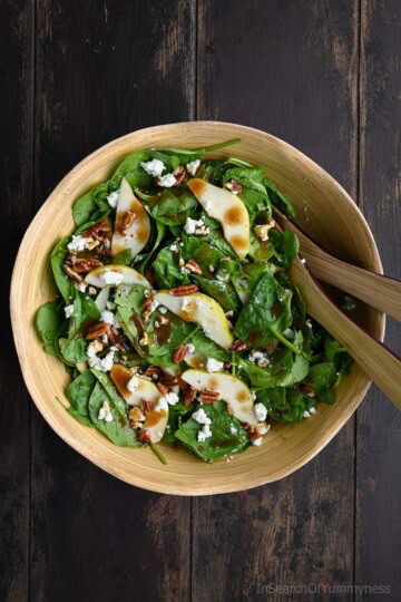 A wooden bowl filled with spinach pear salad topped with crumbled goat cheese, toasted pecans, and balsamic Dijon vinaigrette, with salad tongs resting on the side.