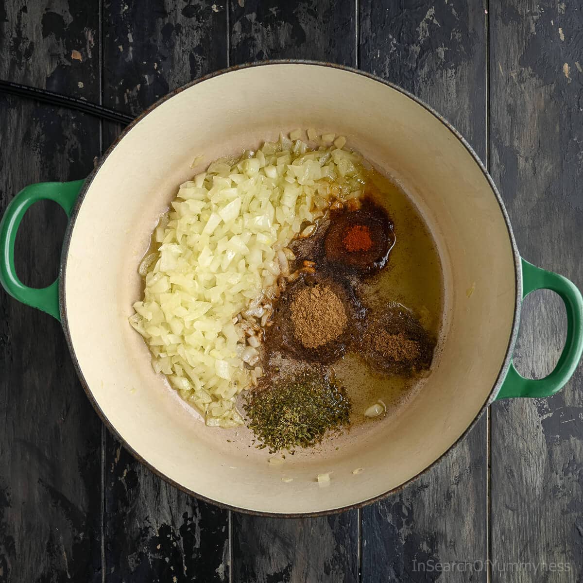 A green Le Creuset French oven with translucent onions and spices toasting in oil.