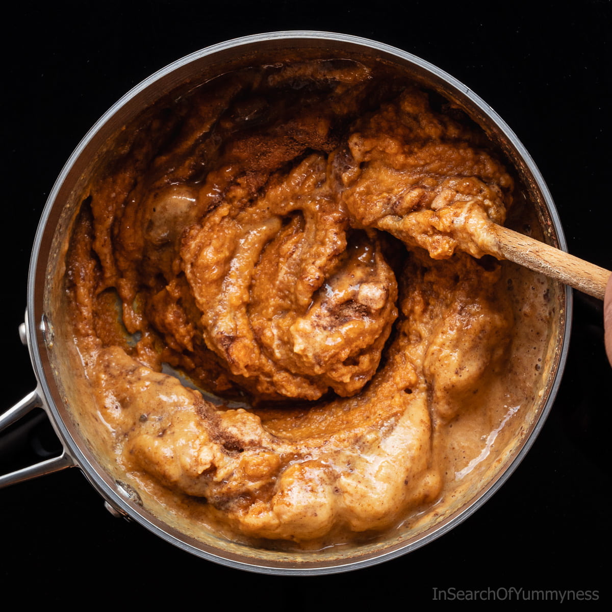 A steel saucepan full of warm pumpkin pie filling ingredients, being stirred with a wooden spoon.
