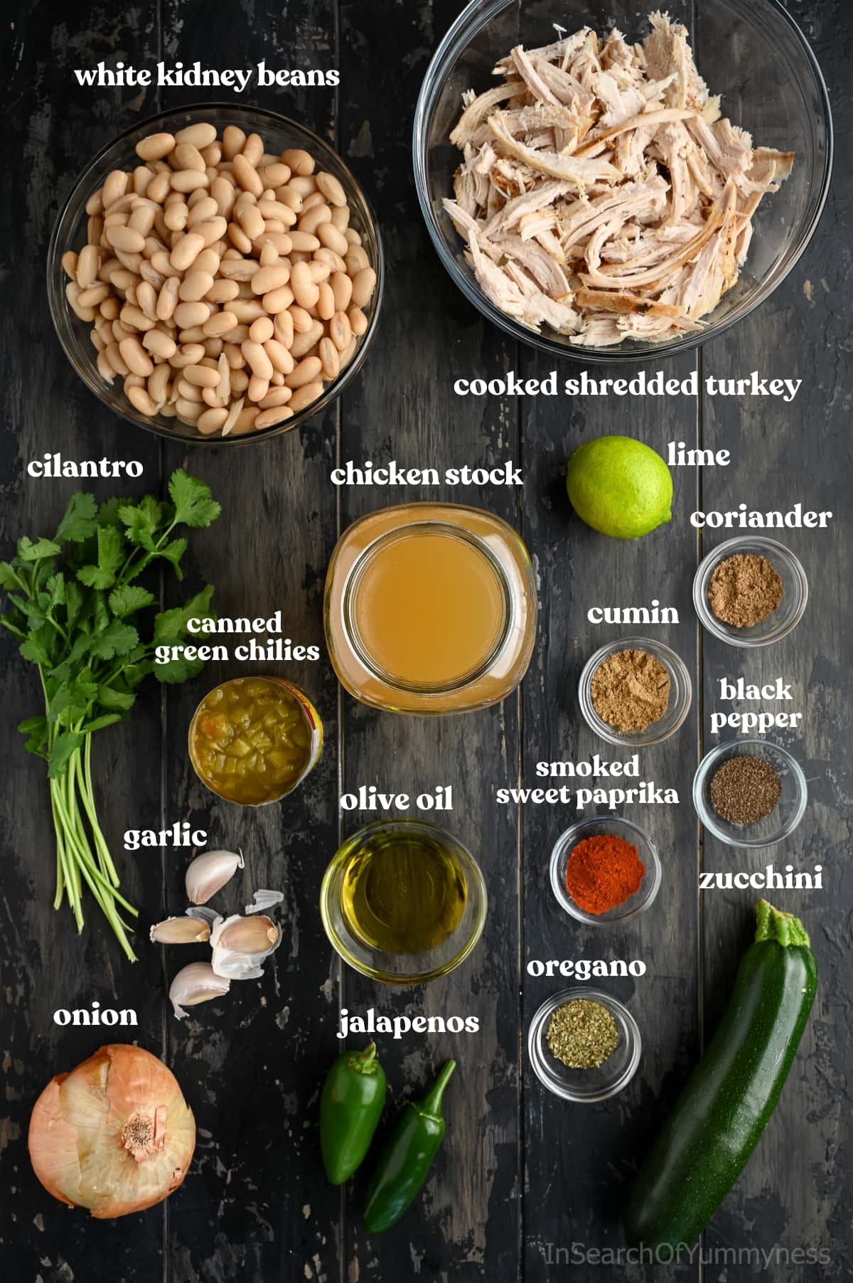 Overhead photo of labeled ingredients for white turkey chili, including shredded turkey, white kidney beans, chicken stock, zucchini, jalapeños, onion, garlic, cilantro, lime, canned green chilies, olive oil, and spices.