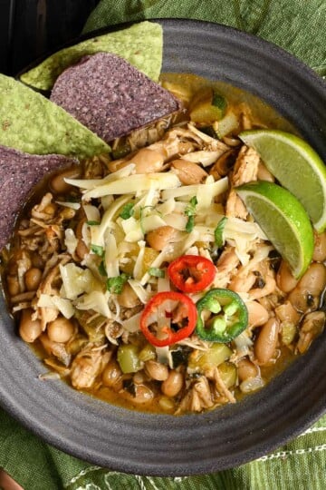 White turkey chili in a dark grey bowl, topped with sliced jalapeños, chopped cilantro, and shredded cheese. Served with tortilla chips and lime wedges.