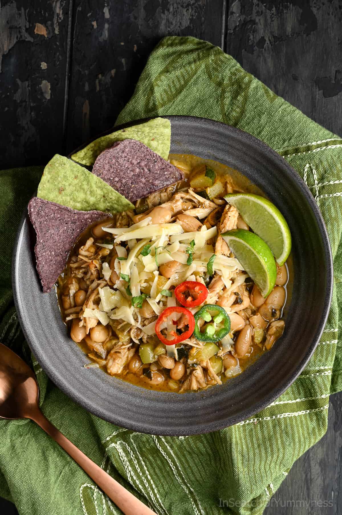 White turkey chili in a dark grey bowl, topped with sliced jalapeños, chopped cilantro, and shredded cheese. Served with tortilla chips and lime wedges.