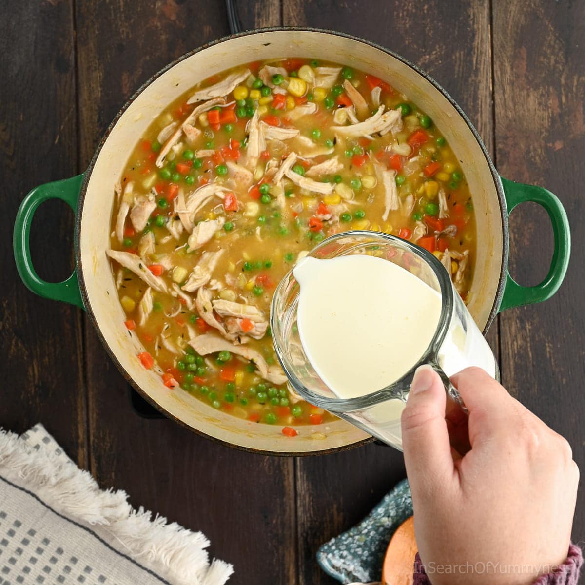 A hand adding heavy cream from a glass measuring jug into the soup pot with the vegetables and chicken.