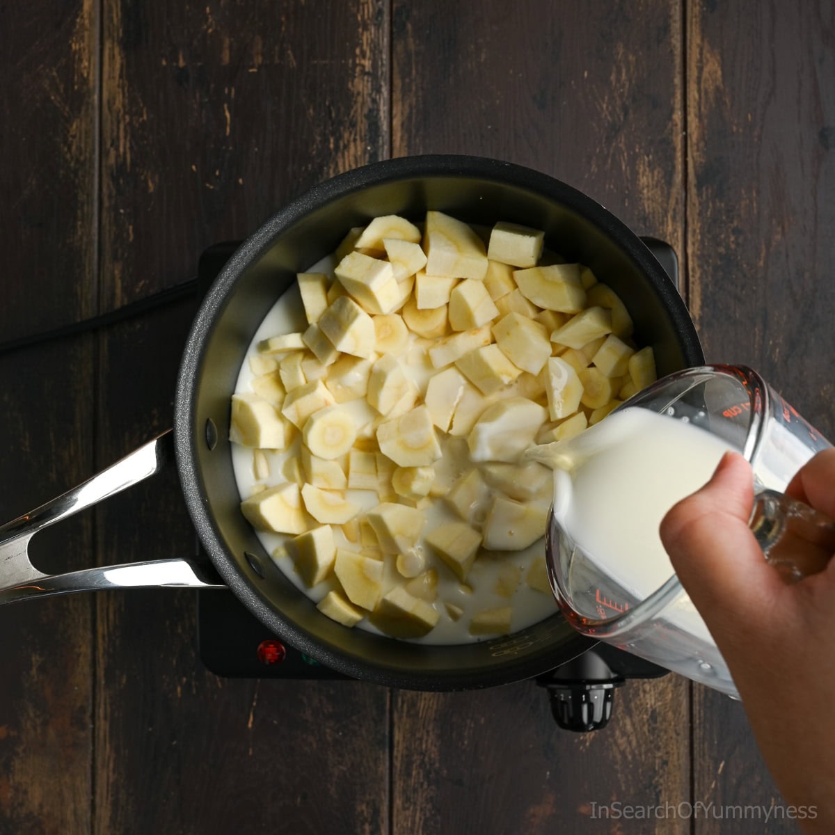 A hand using a glass measuring jug to pour milk into a black non-stick pot containing chopped parsnips.