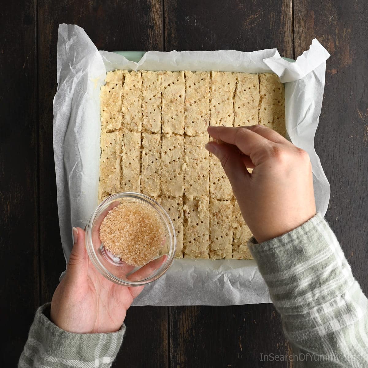 A hand holding a glass bowl containing raw sugar, while the other hand is sprinkling the sugar over unbaked coconut shortbread cookies in a square baking pan lined with parchment paper.