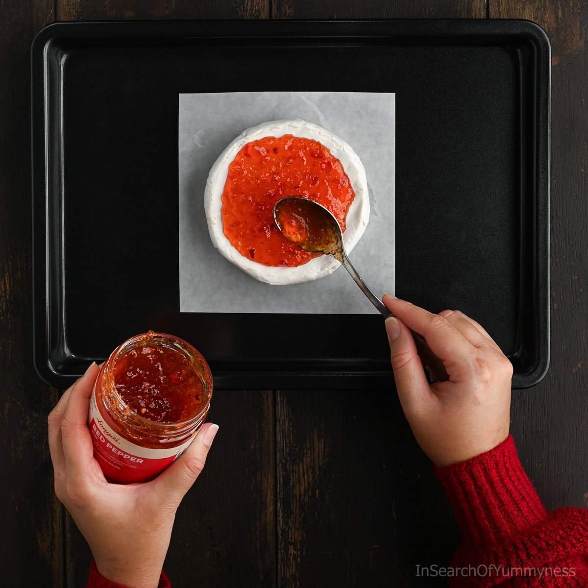 Someone spooning red pepper jelly out of a jar onto an unbaked wheel of brie cheese, which is sitting on a white piece of parchment paper on a black sheet pan.