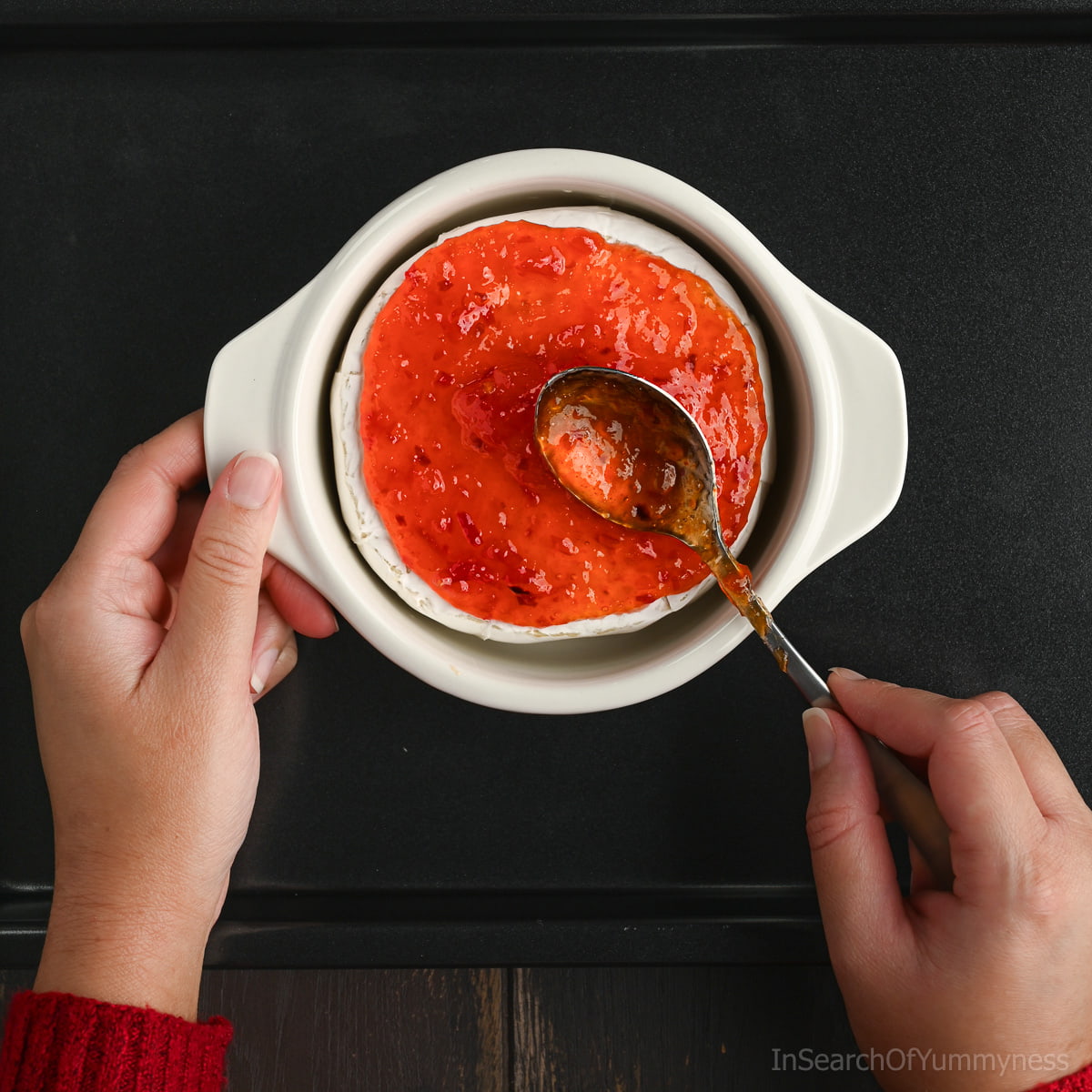 Someone spooning red pepper jelly onto an unbaked wheel of brie cheese in a white brie baker, which is sitting on a black sheet pan.