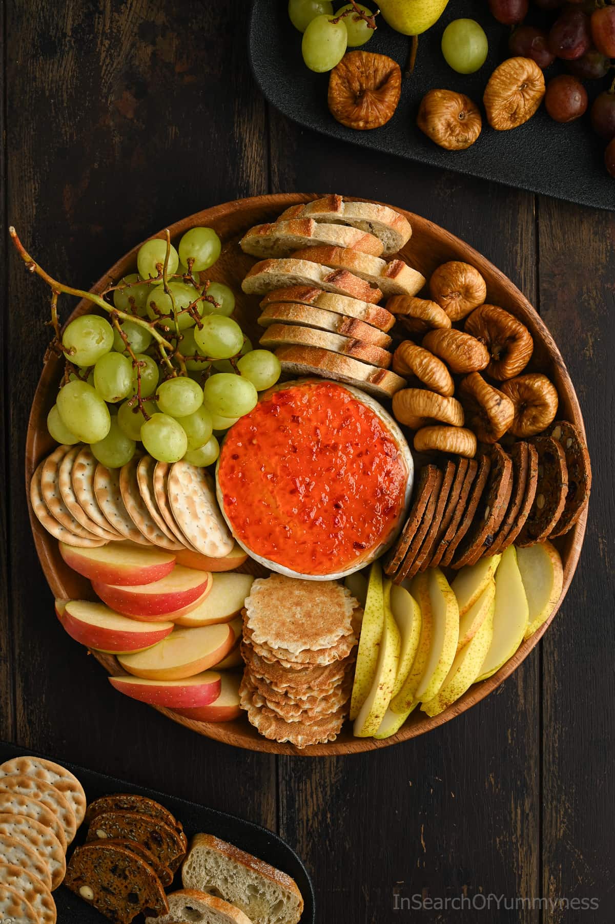 A round brown wooden platter with a wheel of baked Brie topped with red pepper jelly nestled in between crackers, slices of toasted baguette, grapes, apple slices, pear slices and dried figs.