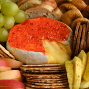 A wheel of baked Brie with melty cheese oozing out, topped with red pepper jelly nestled in between crackers, slices of toasted baguette, grapes, apple slices, pear slices and dried figs.