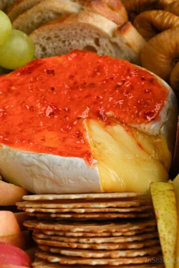 A wheel of baked Brie with melty cheese oozing out, topped with red pepper jelly nestled in between crackers, slices of toasted baguette, grapes, apple slices, pear slices and dried figs.