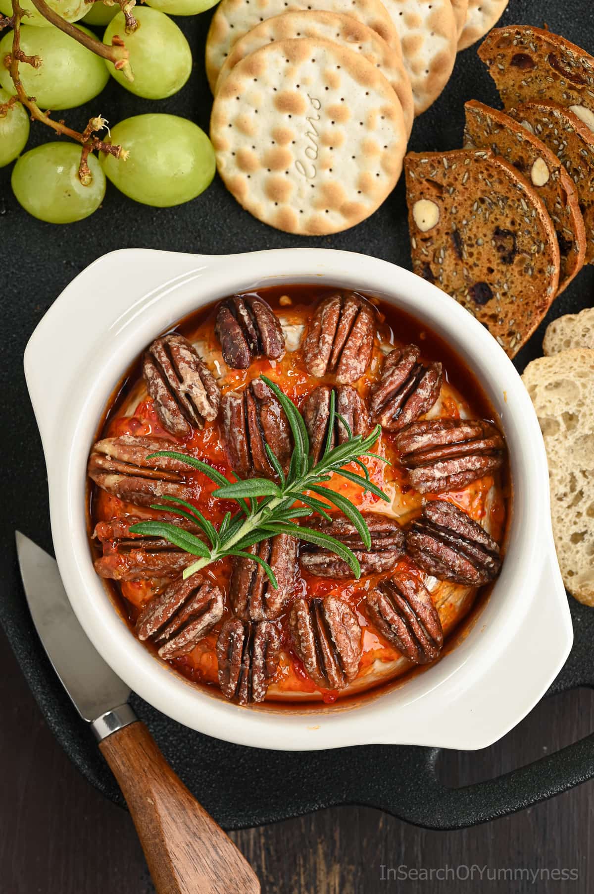 A wheel of baked Brie in a white Brie baker, topped with red pepper jelly, candied pecans and a spring of rosemary. The brie baker is on a tray with grapes and various crackers.