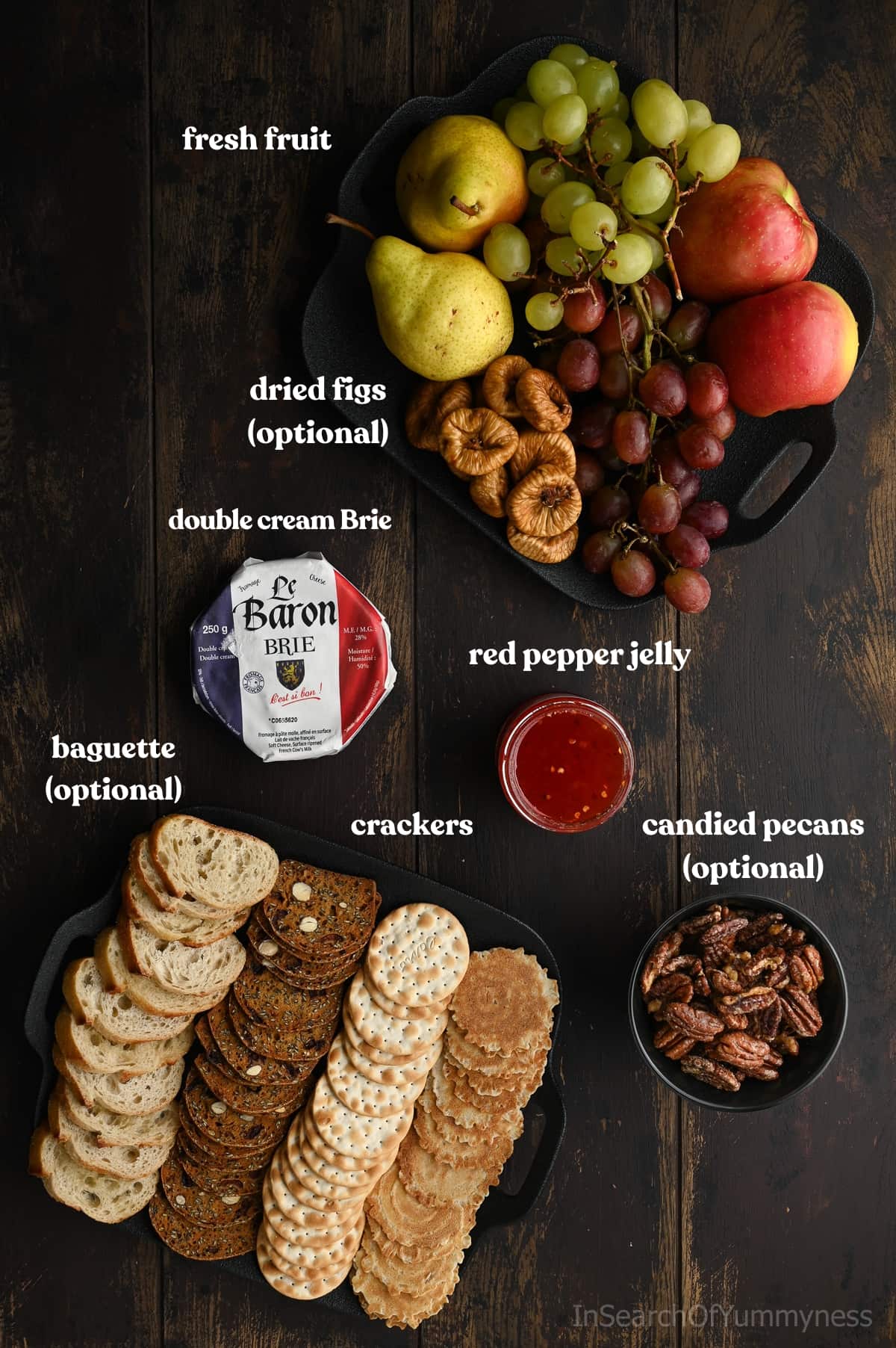 Ingredients for baked brie with red pepper jelly. A small wheel of double cream Brie, a platter with fresh fruit, a platter with crackers and slices of baguette, a jar of red pepper jelly and a bowl of candied pecans arranged on a dark wood surface.
