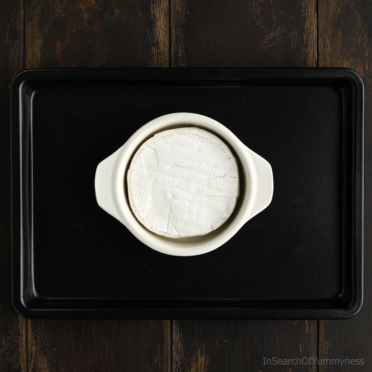 A wheel of Brie cheese inside a white brie baker on a black sheet pan, waiting to go into the oven.