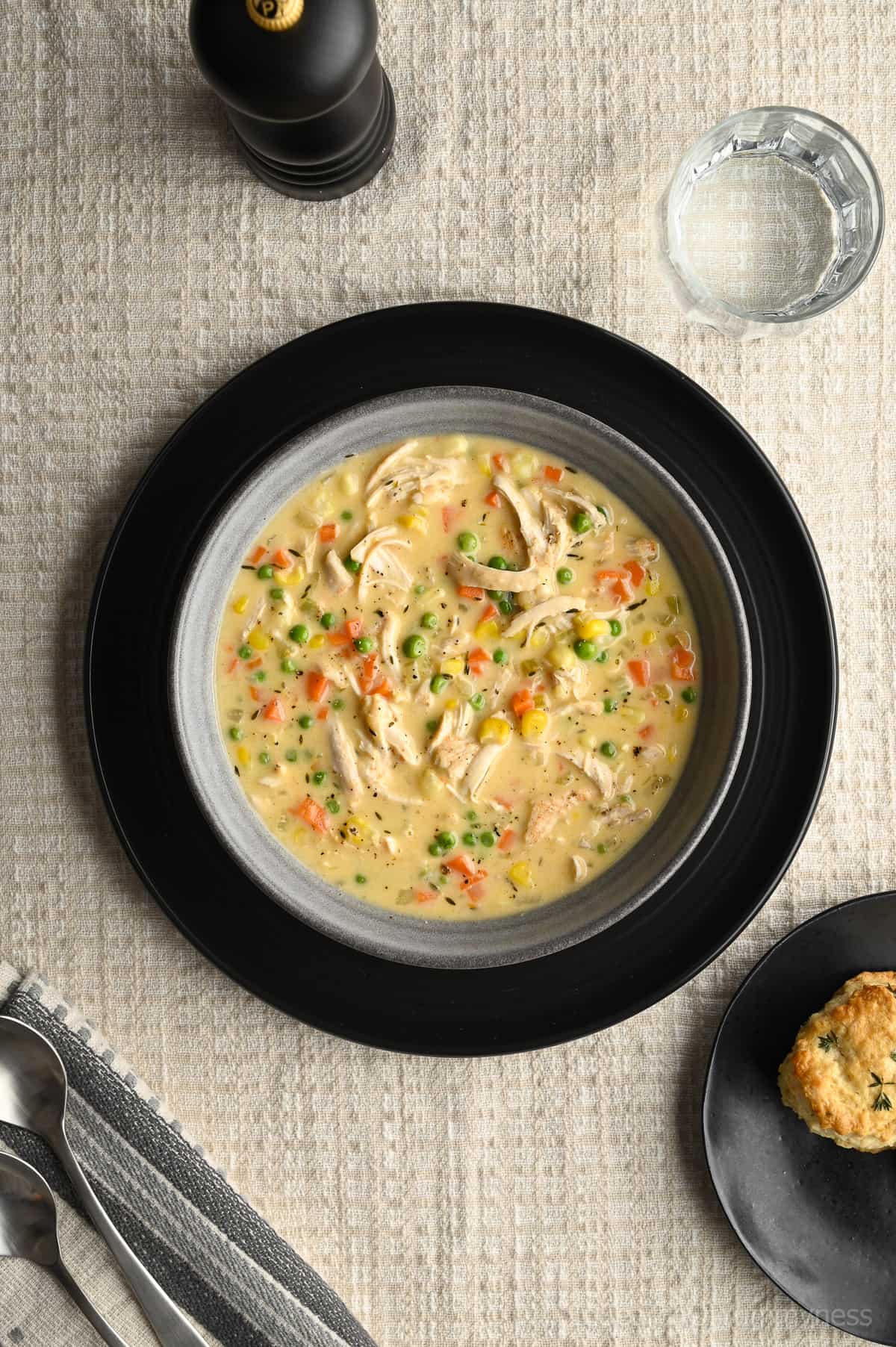 A bowl of chicken pot pie soup in a grey bowl on a black charger plate. Surrounded by a black pepper mill, a glass of water, a black plate with a biscuit and a towel with two silver spoons.