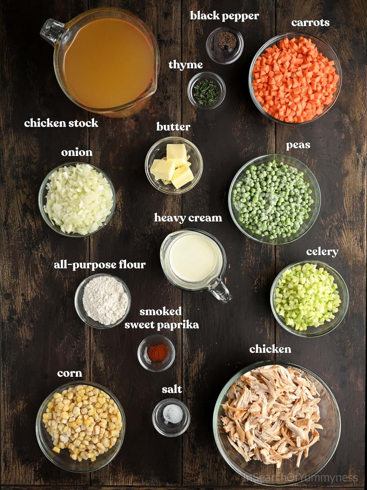 Ingredients for chicken pot pie soup. Bowls of butter, spices, vegetables, and chicken are next to measuring jugs full of stock and cream, arranged on a dark wood surface.