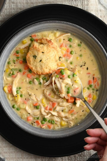 Someone wearing a purple sweater using a spoon to lift a spoonful of warm chicken pot pie soup out of a grey bowl. The soup has shredded chicken, diced carrots, diced celery, peas and corn kernels in it. There is also half of a biscuit sitting on top of the soup.