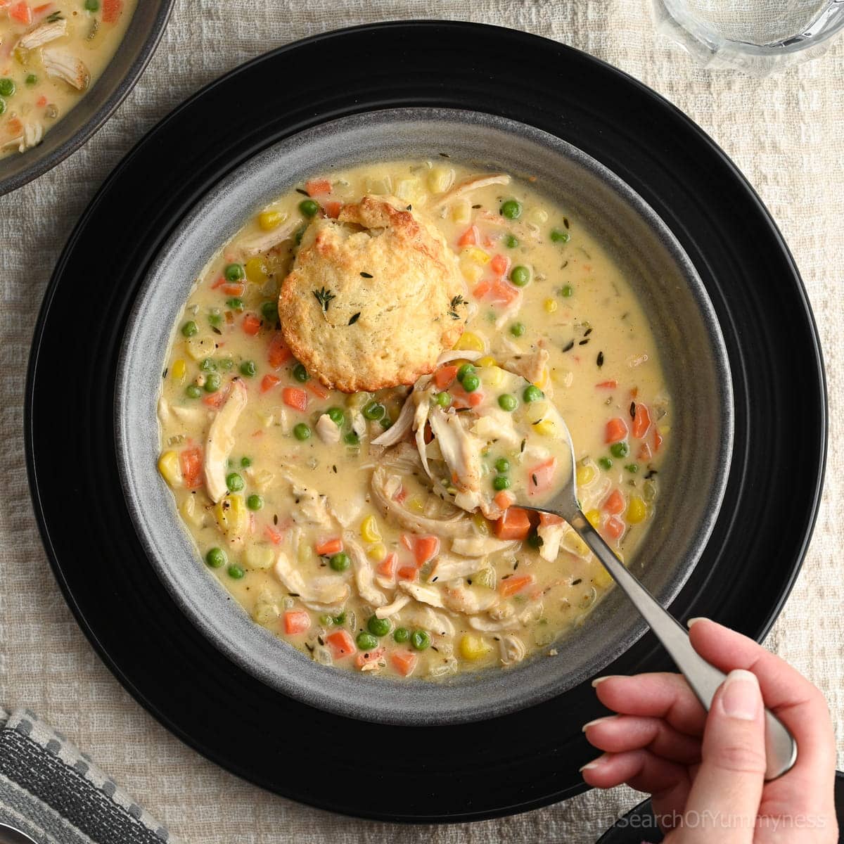Someone wearing a purple sweater using a spoon to lift a spoonful of warm chicken pot pie soup out of a grey bowl. The soup has shredded chicken, diced carrots, diced celery, peas and corn kernels in it. There is also half of a biscuit sitting on top of the soup.