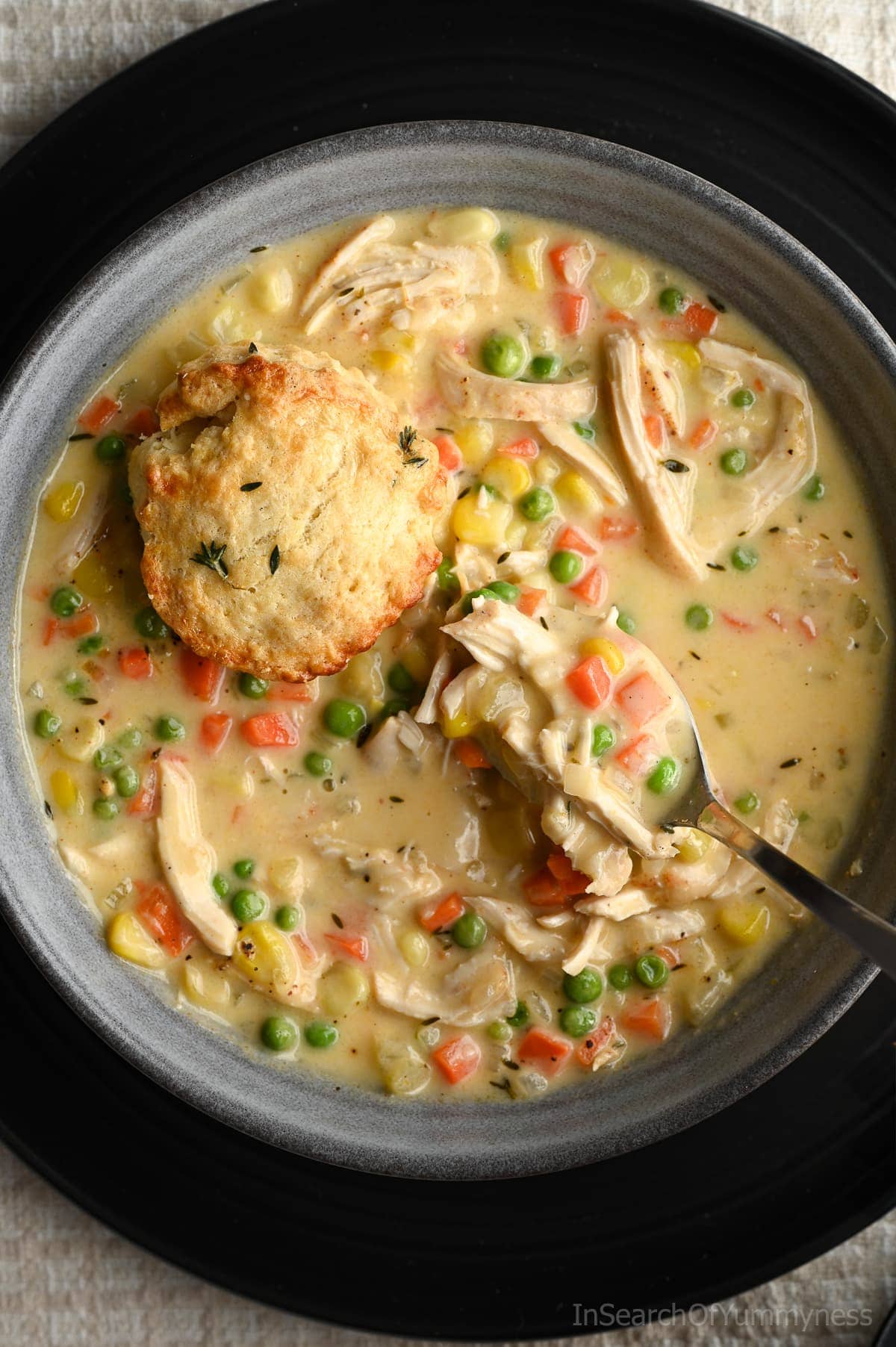 A spoon lifting a spoonful of chicken pot pie soup out of a bowl, showing off the shredded chicken and vegetables. There is also half of a fresh biscuit on top of the soup.