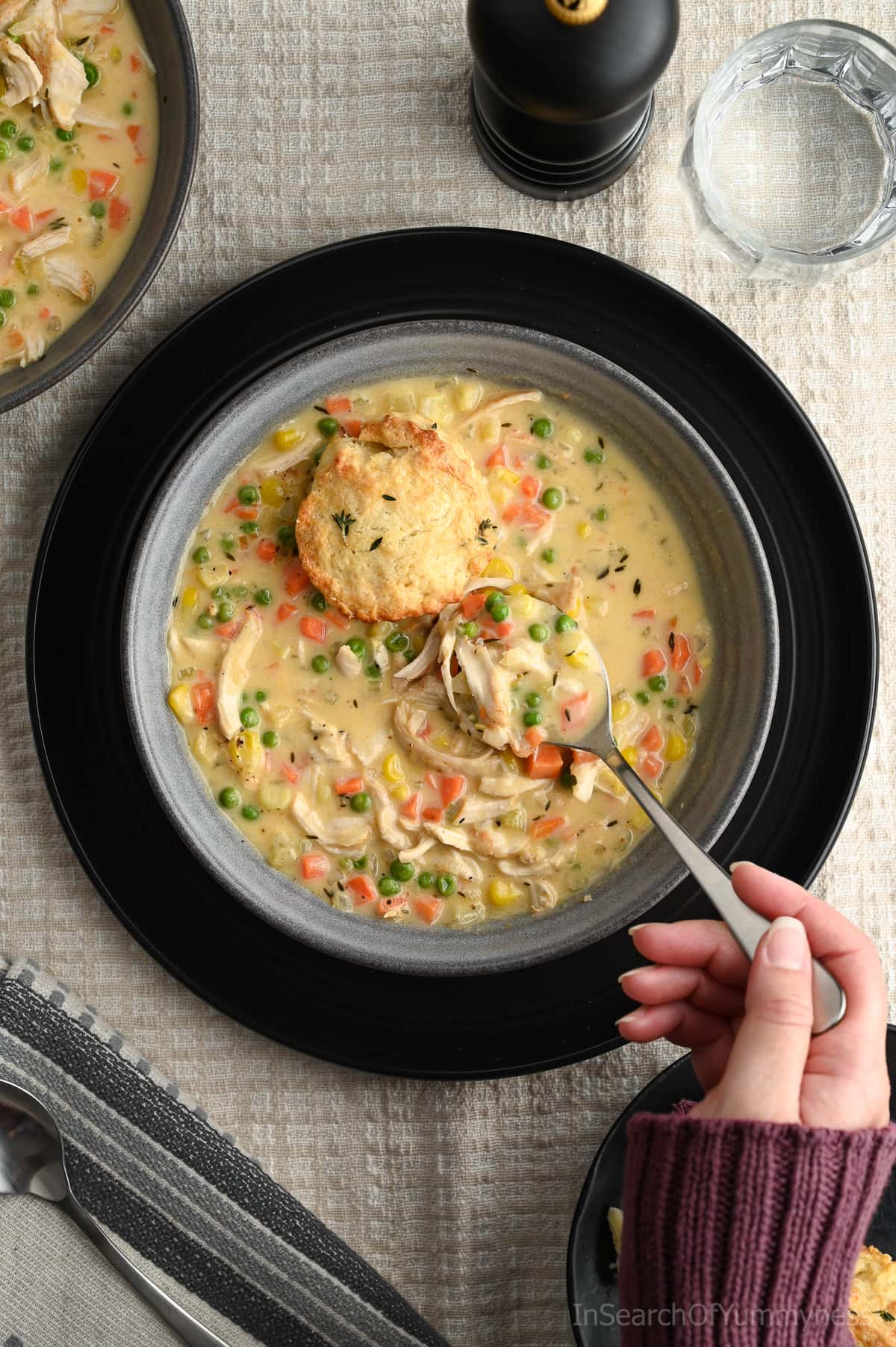 Someone wearing a purple sweater using a spoon to lift a spoonful of warm chicken pot pie soup out of a grey bowl. The soup has shredded chicken, diced carrots, diced celery, peas and corn kernels in it. There is also half of a biscuit sitting on top of the soup.