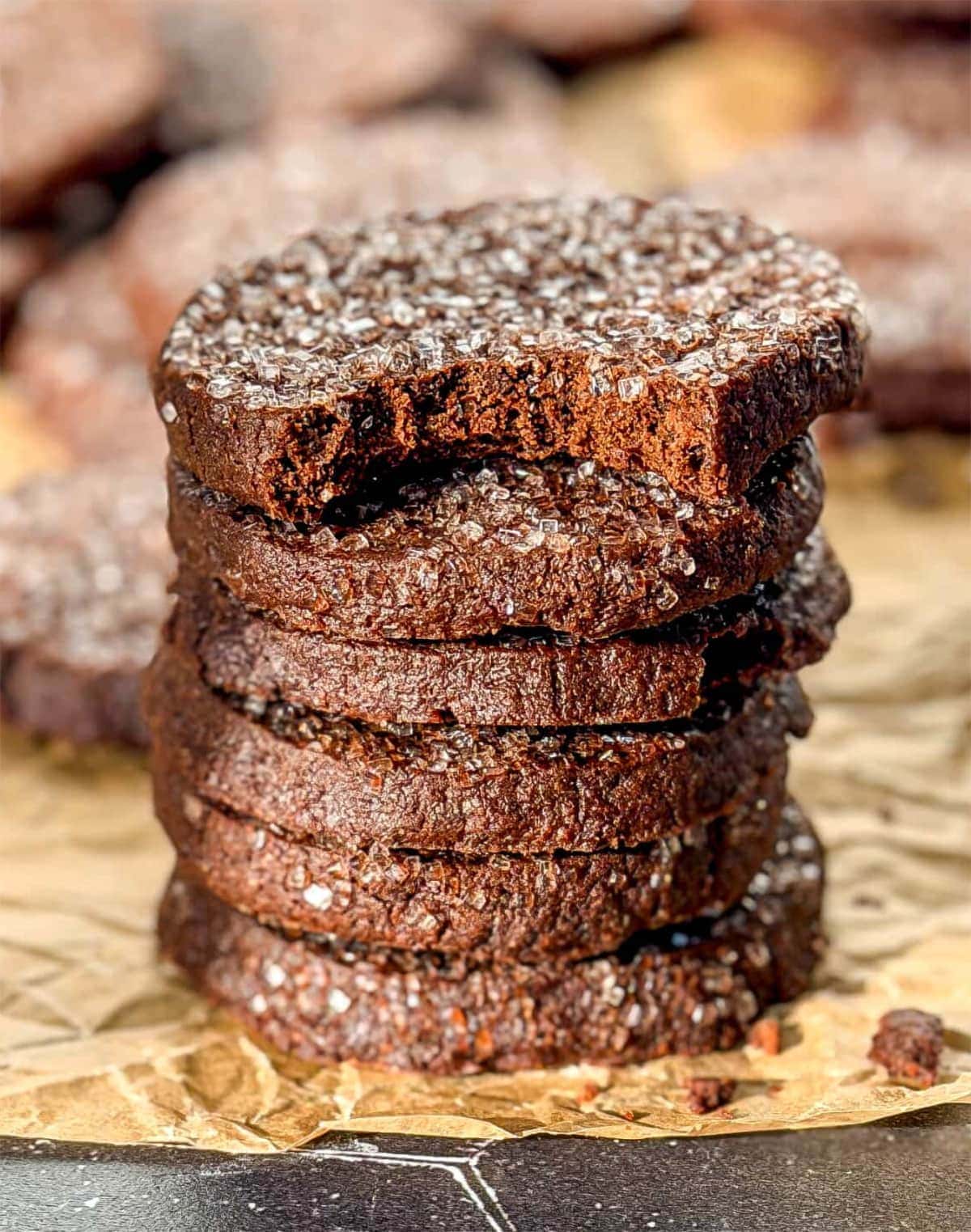 A stack of 6 chocolate shortbread cookies on parchment paper. The top cookie has a bite taken out of it. Photo by Cynthia from But First We Brunch.