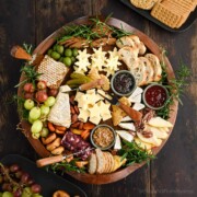 A large round cheeseboard packed with 5 different kinds of cheese, a square of fresh honeycomb, fresh apple slices, grapes and pear slices, and various condiments.