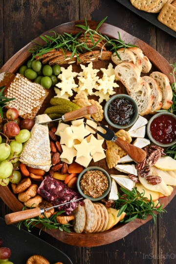 A large round cheeseboard packed with 5 different kinds of cheese, a square of fresh honeycomb, fresh apple slices, grapes and pear slices, and various condiments.