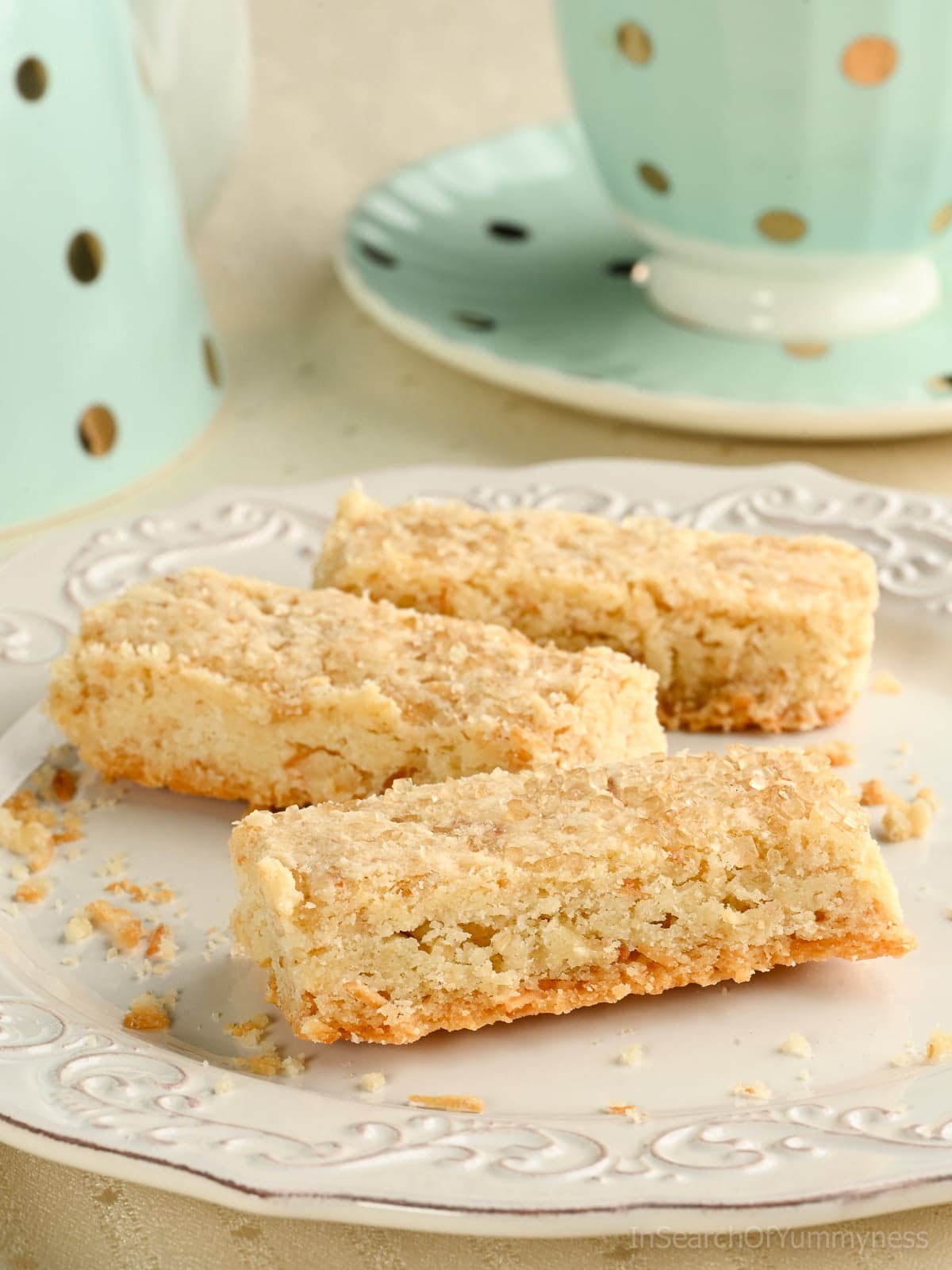 Three coconut shortbread cookies made with toasted coconut on a white plate, with a mint green polka dot teacup in the background.