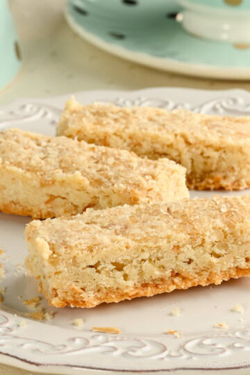 Three coconut shortbread cookies made with toasted coconut on a white plate, with a mint green polka dot teacup in the background.