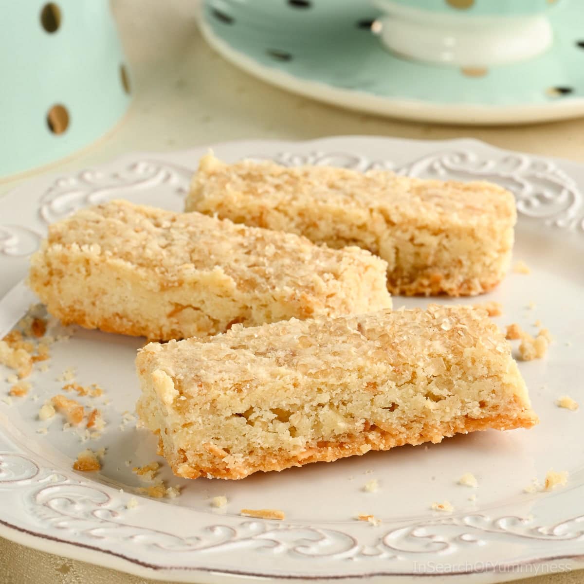 Three coconut shortbread cookies made with toasted coconut on a white plate, with a mint green polka dot teacup in the background.