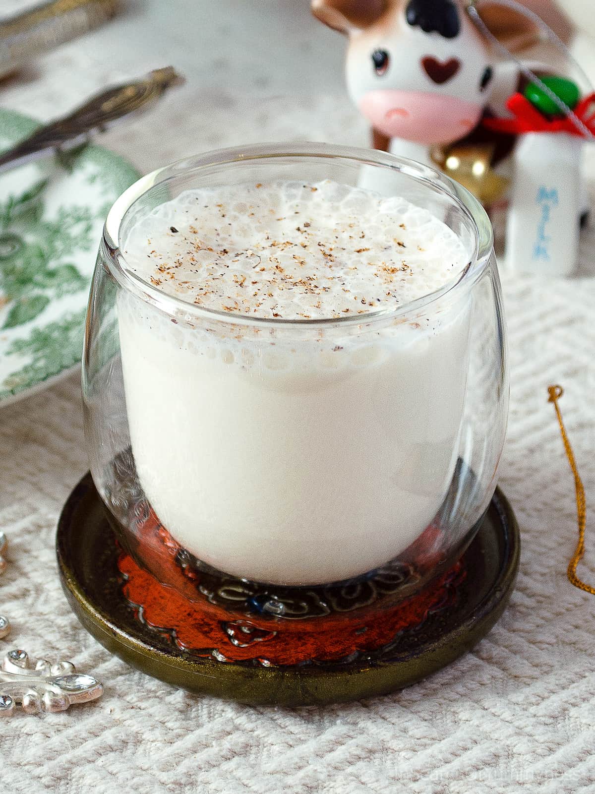 A clear double-walled glass full of non-alcoholic eggless eggnog, sitting on a fancy coaster on top of a white tablecloth.