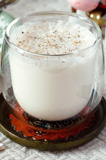 A clear double-walled glass full of non-alcoholic eggless eggnog, sitting on a fancy coaster on top of a white tablecloth.