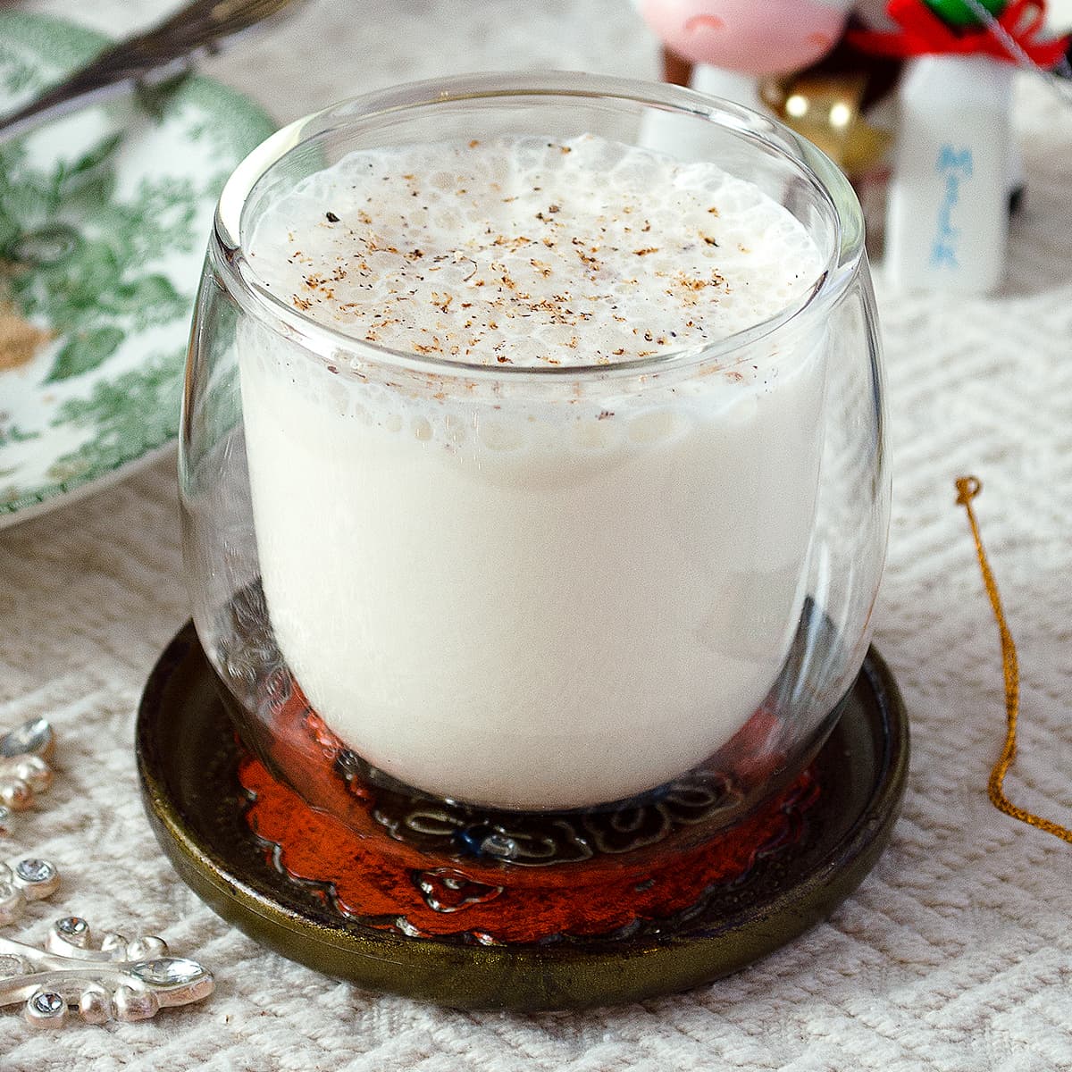 A clear double-walled glass full of non-alcoholic eggless eggnog, sitting on a fancy coaster on top of a white tablecloth.