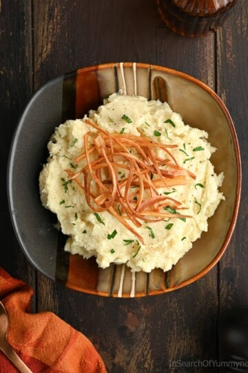 Mashed parsnips in a ceramic bowl, topped with crispy parsnip strips and chopped parsley, set on a dark wooden table.