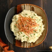 Mashed parsnips in a ceramic bowl, topped with crispy parsnip strips and chopped parsley, set on a dark wooden table.