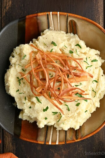 Mashed parsnips in a ceramic bowl, topped with crispy parsnip strips and chopped parsley, set on a dark wooden table.
