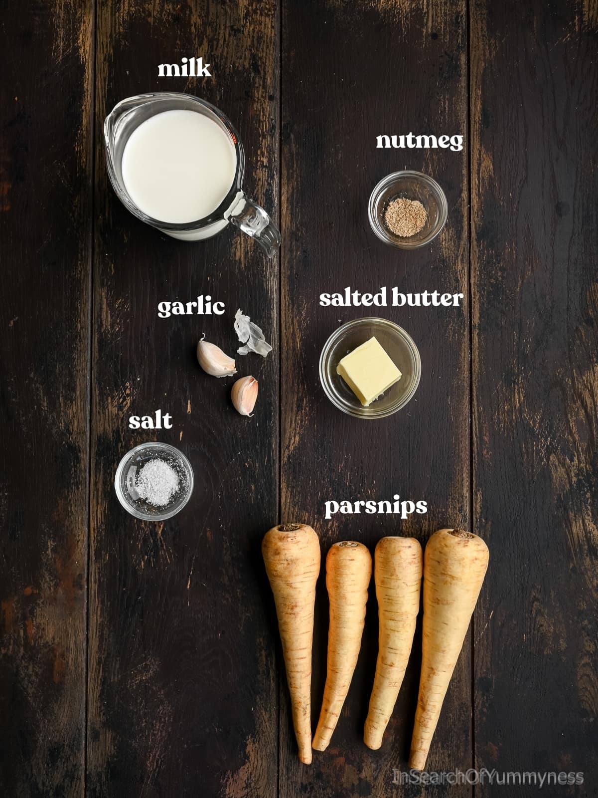Overhead view of ingredients for mashed parsnips, including parsnips, milk, butter, garlic, salt, and nutmeg arranged on a dark wooden surface.