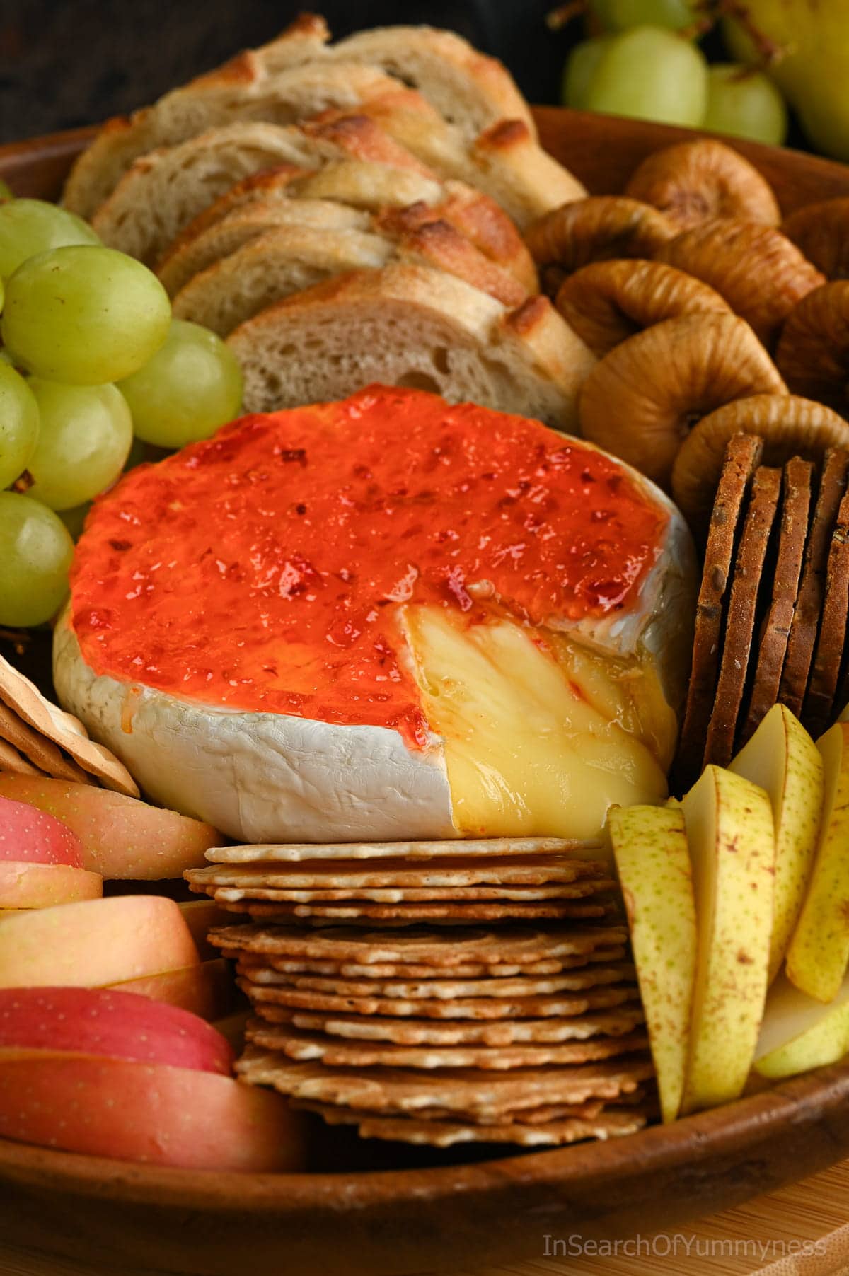 A wheel of baked Brie with melty cheese oozing out, topped with red pepper jelly nestled in between crackers, slices of toasted baguette, grapes, apple slices, pear slices and dried figs.
