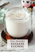 A clear double-walled glass full of non-alcoholic eggless eggnog, sitting on a fancy coaster on top of a white tablecloth. Text overlay says "Non-Alcoholic Eggless Eggnog".