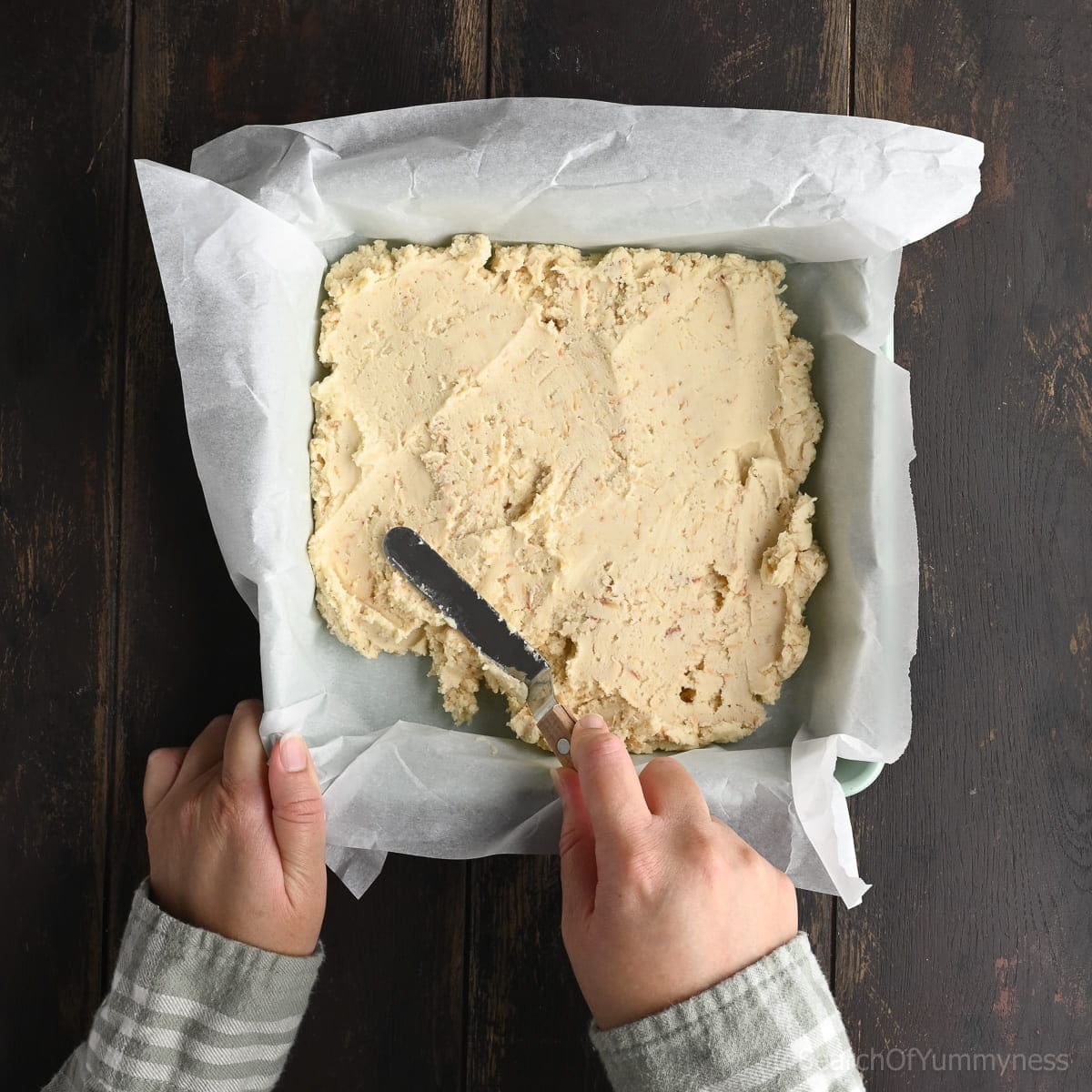 A hand holding a square baking pan lined with parchment while another hand uses a small palette knife to evenly spread out the coconut shortbread dough.