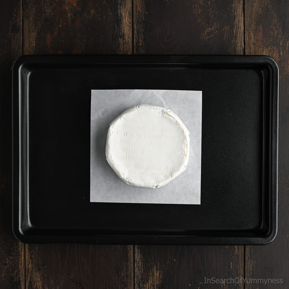 A wheel of Brie cheese sitting on a square piece of parchment paper on a black sheet pan, waiting to go into the oven.